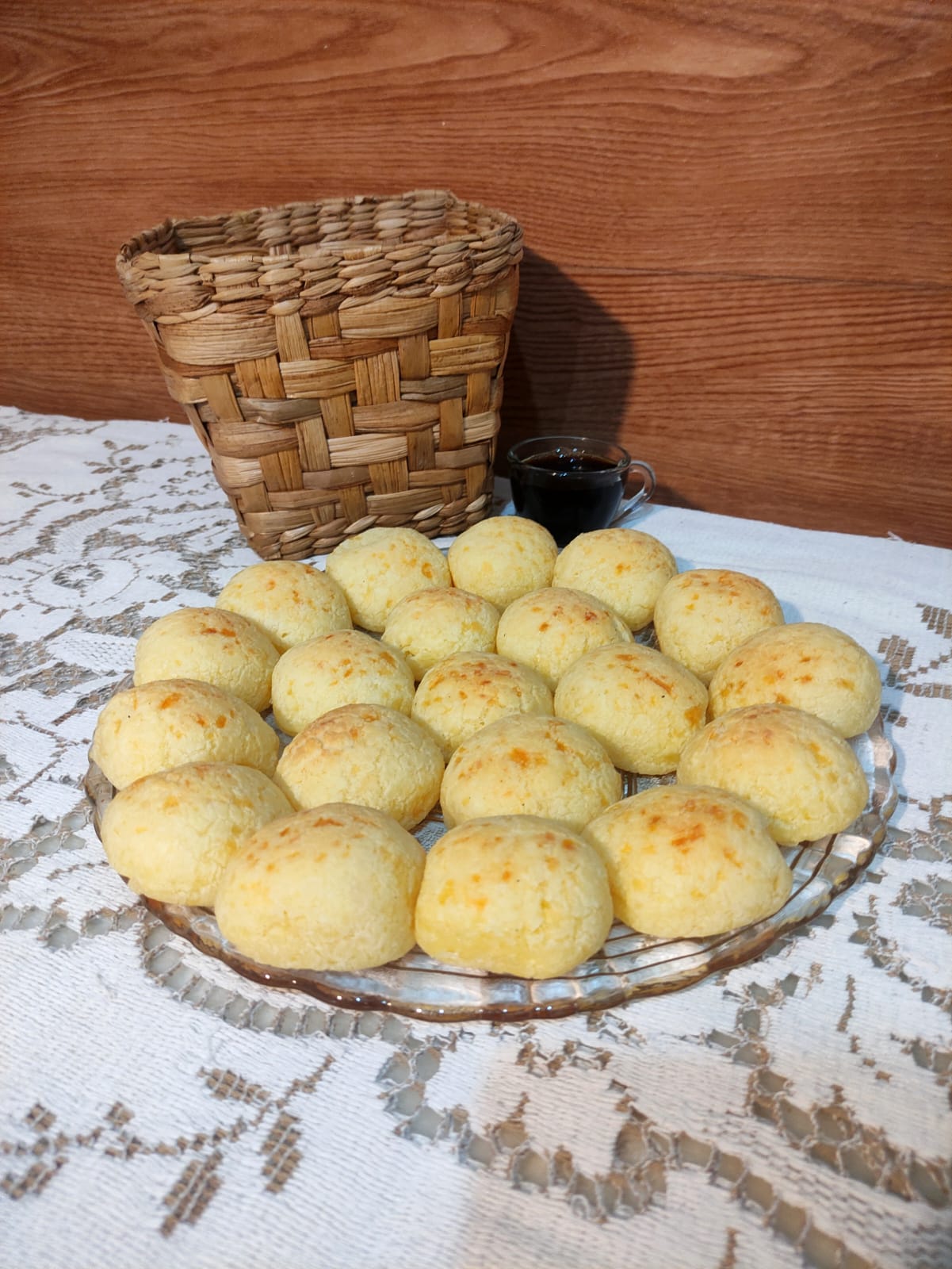 Travessa de pão de queijo quentinho ao lado de cesta de palha e uma xícara de café